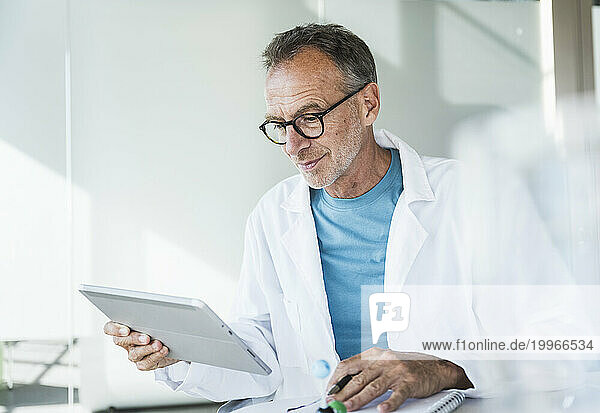 Smiling doctor using tablet PC and sitting with note pad at desk