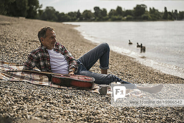 Contemplative senior man with guitar sitting on blanket at beach