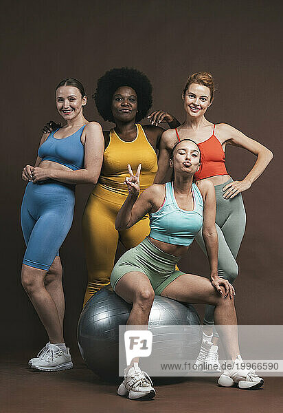 Smiling multiracial female friends with fitness ball against brown background