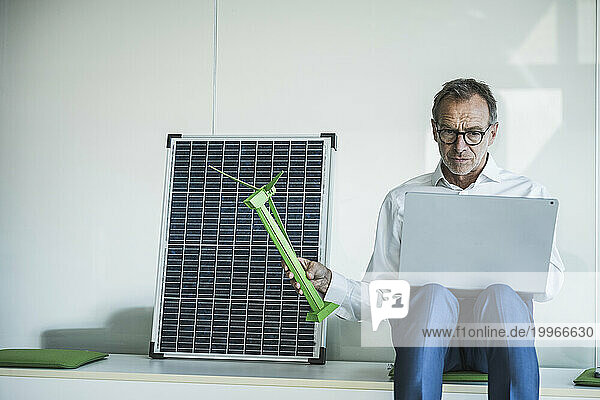 Senior businessman using laptop and holding wind turbine model near solar panel in office