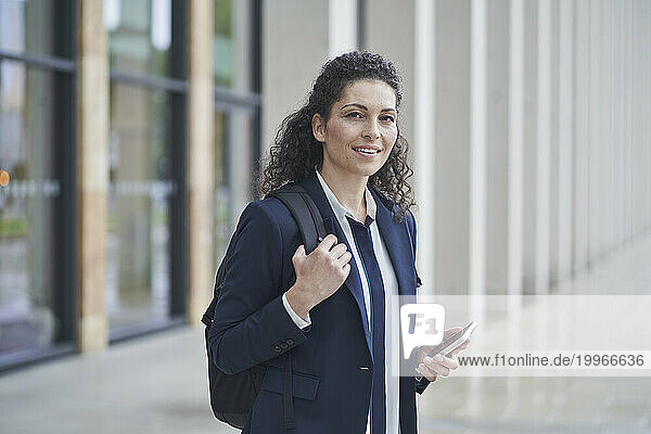 Smiling businesswoman holding smart phone near building