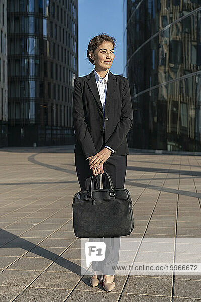 Smiling businesswoman holding briefcase and standing on pavement at sunset