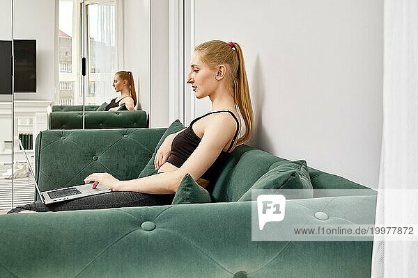 Aktive Frau in Sportkleidung  die auf dem Sofa sitzt und online eine Verbindung mit einem Trainer herstellt