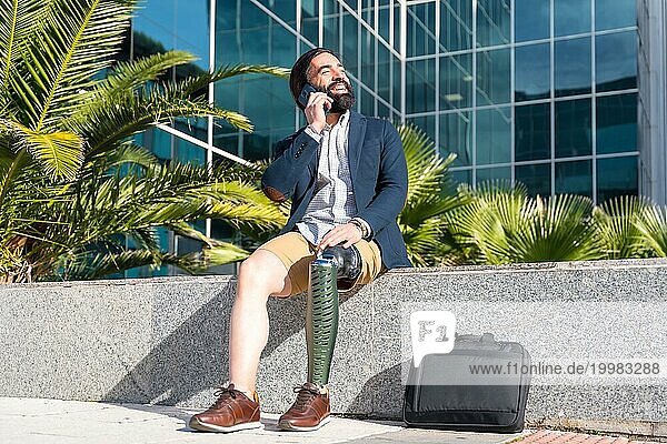Geschäftsmann mit Beinprothese  der lächelnd mit dem Handy spricht  das vor einem Finanzgebäude steht