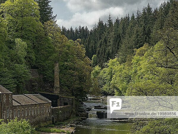 Ein ruhiger Fluss fließt durch einen dichten Wald mit alten Gebäuden am Ufer  Ehemaliges Maschinenhaus am Fluss Clyde. Unesco- Weltkulturerbe New Lanark -England  Großbritannien  Europa