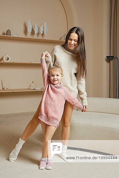 Eine fröhliche Mutter und ihre kleine Tochter tanzen zusammen in einem gemütlichen Wohnzimmer. Die Tochter trägt einen rosa Pullover  während die Mutter einen grauen Pullover trägt