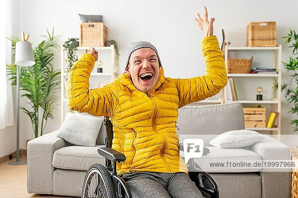 Porträt eines glücklichen behinderten Mannes im Rollstuhl  der vor Freude die Hände hebt und im Wohnzimmer zu Hause feiert