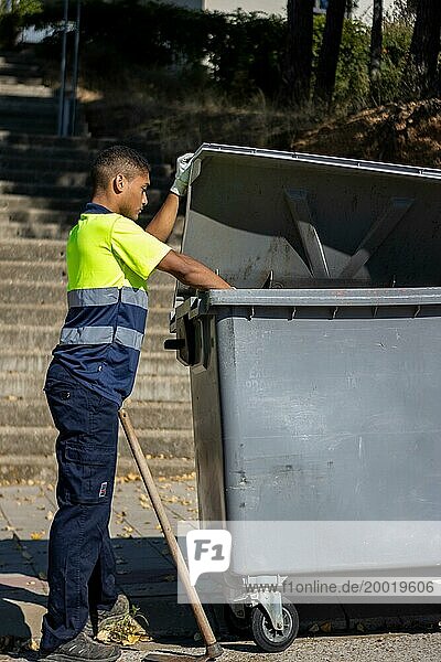 Vertikale Ansicht eines städtischen Arbeiters in Uniform  der einen Müllsack in einen großen Müllcontainer wirft  während er den Deckel des Containers hochhält