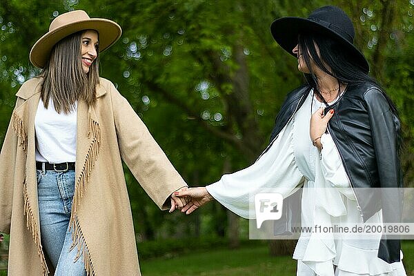 Vorderansicht eines stilvollen lesbischen Paares mit Hut  das sich an den Händen hält und im Park spazieren geht  während es sich gegenseitig anschaut
