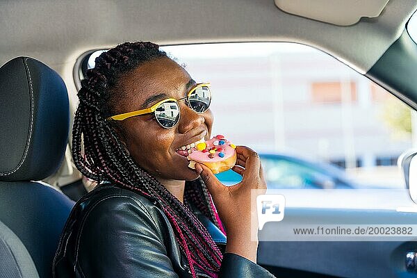 Bezaubernde afrikanische Frau lächelt  während sie in einem Auto sitzend einen Doughnut isst