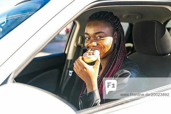Blick von außen auf eine fröhliche afrikanische Frau  die in einen Donut beißt  während sie im Haus sitzt