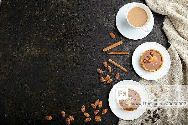 Süße Törtchen mit Mandeln und Karamellcreme mit einer Tasse Kaffee auf schwarzem Betonhintergrund und Leinentextil. Draufsicht  flat lay  copy space