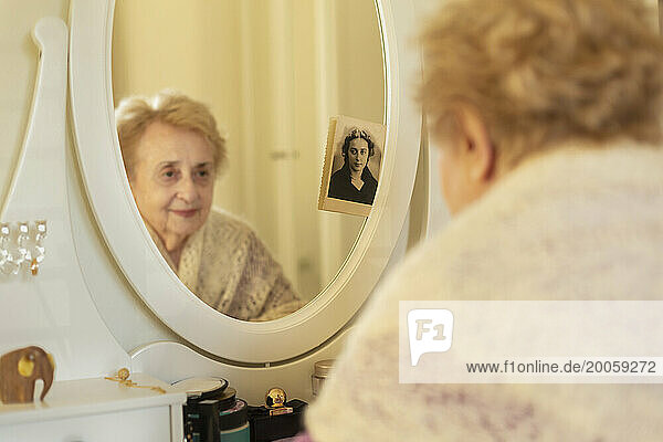 Smiling senior woman looking at reflection in mirror next to old photograph
