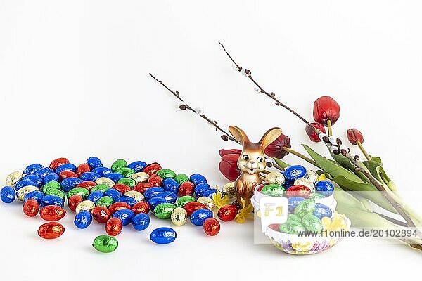 Eine Ansammlung von bunten Schokoladeneiern und einem Osterhasen neben Tulpen und Palmkätzchen  weißer Hintergr
