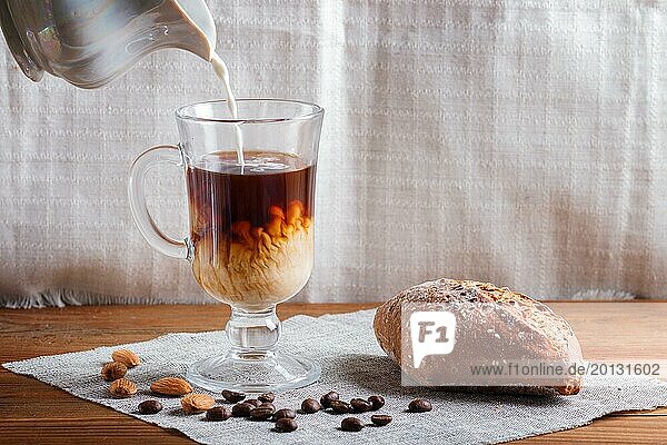 Glass cup of coffee with cream poured over and bun on a wooden background and linen textile. close up  copy space