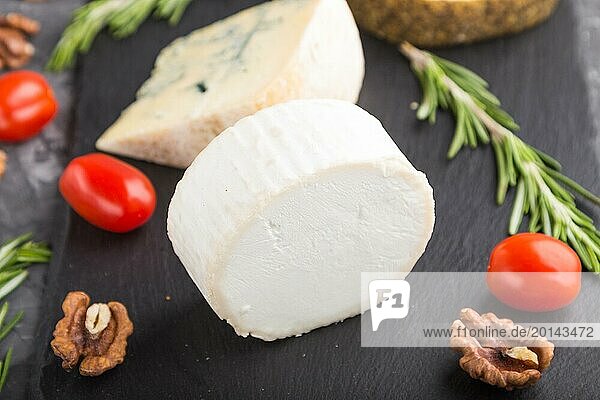 White goat cheese and various types of cheese with rosemary and tomatoes on black slate board on a black concrete background. Side view  close up  selective focus
