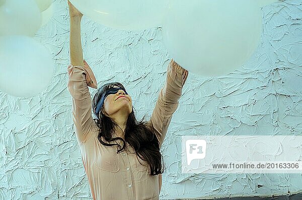 Eine spielerische junge erwachsene hispanische Frau mit verbundenen Augen streckt sich in einer heiteren Geste zwischen zwei großen Luftballons in die Höhe  künstlerische Performance  spielerischer Tanz