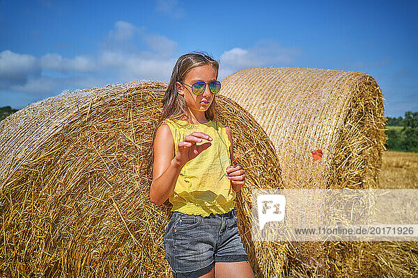 Girl wearing sunglasses and leaning on bale of straw in field
