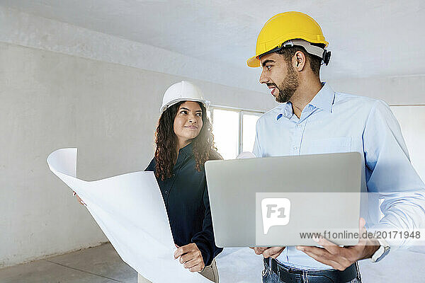 Smiling architects discussing with blueprint and laptop at site