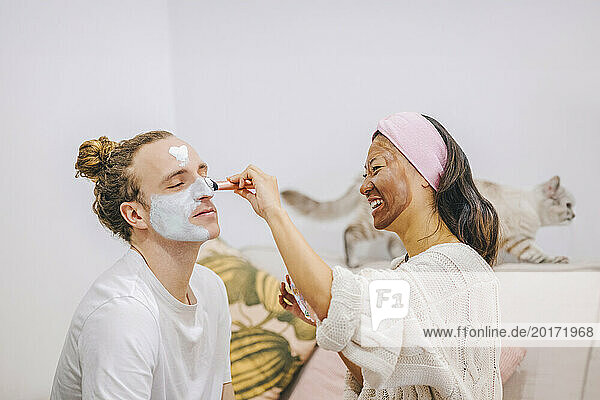 Happy woman applying skincare mask on boyfriend's face at home