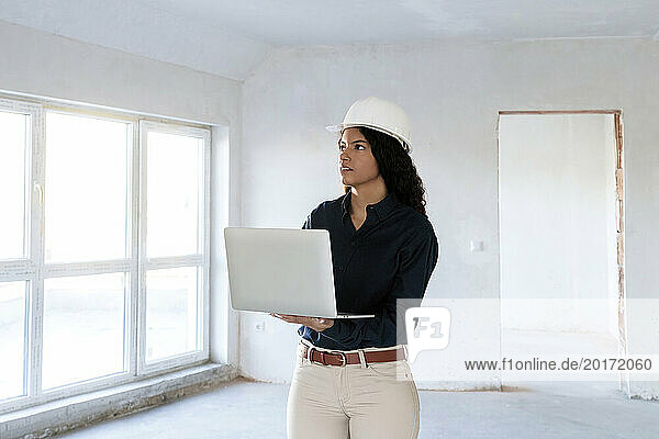 Architect wearing hardhat and standing with laptop at site