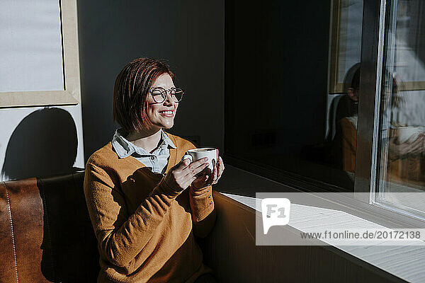 Lächelnde Frau hält Kaffeetasse in der Hand und genießt Sonnenlicht im Café