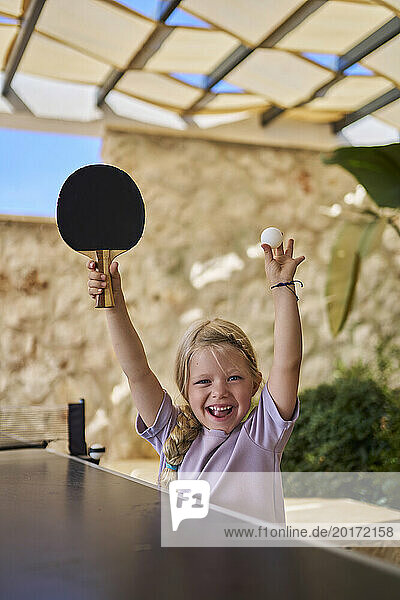 Happy girl cheering with table tennis racket and ball at villa