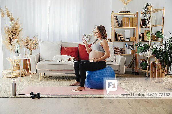 Pregnant woman using smart phone sitting on fitness ball in living room at home