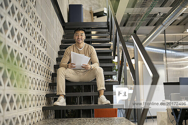 Smiling young businessman with documents sitting on staircase at office