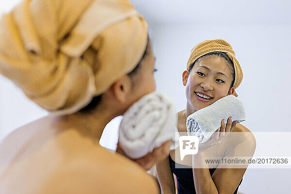 Smiling young woman wiping face with towel in bathroom at home