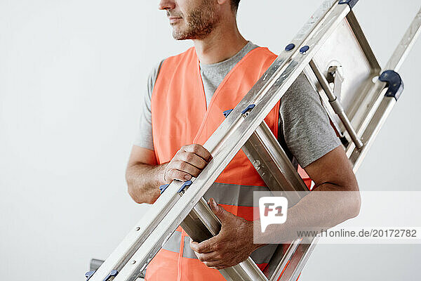 Mature technician carrying ladder in front of wall at home