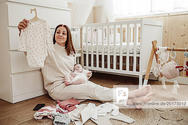 Woman holding daughter and choosing baby clothes near crib