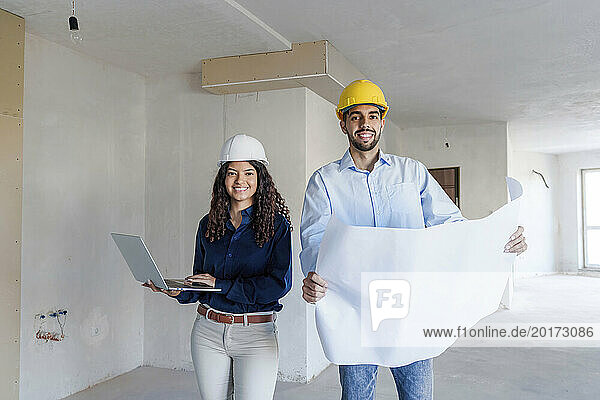 Smiling architects standing with laptop and blueprint at site