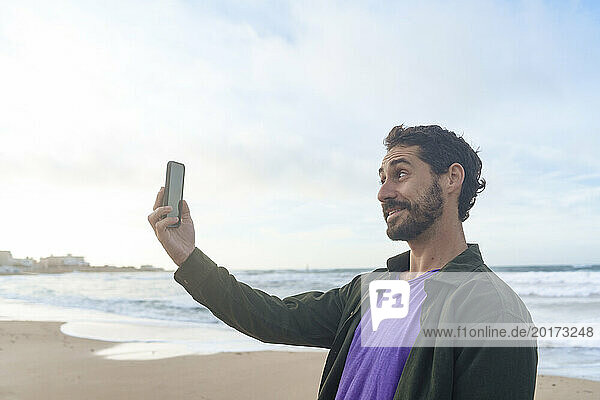Smiling man taking selfie through smart phone in front of sea at beach