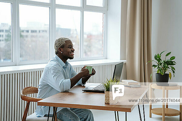 Smiling freelancer sitting with coffee cup and laptop at desk