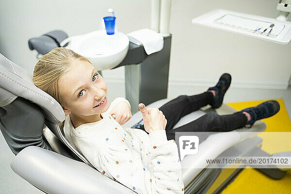 Smiling girl showing thumbs up gesture sitting at dentist's office