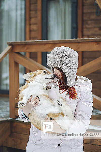 Young woman holding Corgi dog in front of log cabin
