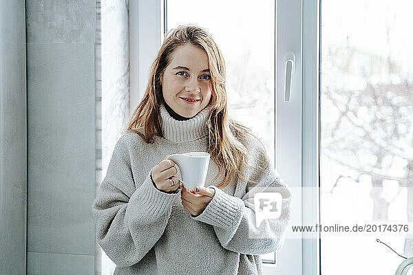 Smiling woman with brown hair holding coffee cup near window at home