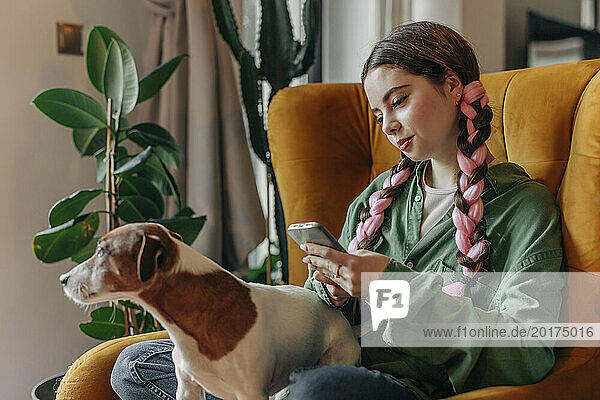 Smiling young woman sitting with dog and using smart phone at home
