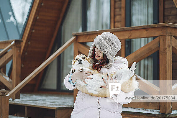 Smiling young woman holding Corgi dog in front of log cabin