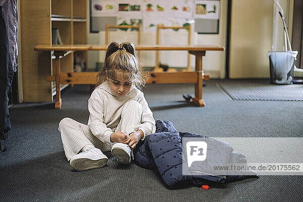 Girl tying shoelace while sitting with jacket on floor in classroom at kindergarten