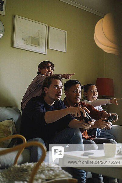 Serious gay men playing video game with daughters pointing at home