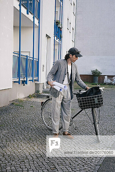 Full length of male architect holding blueprint rolls standing by bicycle on footpath