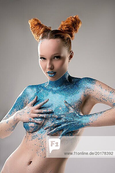 Selbstbewusstes verführerisches junges weibliches Modell mit kreativer Frisur und blauem Glitzer auf den Lippen und nacktem Körper  der die Brust bedeckt und in die Kamera schaut  vor grauem Hintergrund