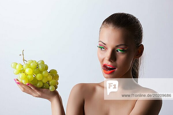 Lustige junge Frau schaut auf grüne Trauben mit Glow Make up
