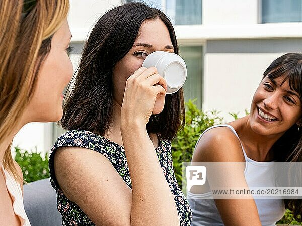 Drei Frauen sitzen draußen und eine von ihnen trinkt aus einer weißen Tasse  während sie in die Kamera schaut. Sie lächeln alle und scheinen die Gesellschaft des anderen zu genießen