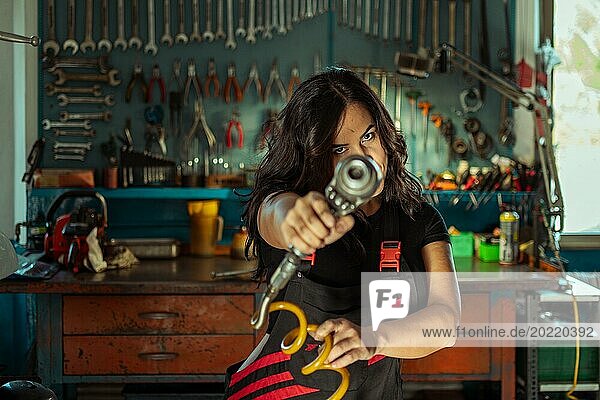 Intensive sexy Frau Mechanikerin zielt ein Werkzeug auf die Kamera in einer Werkstatt  eine komplette Werkzeugtafel im Hintergrund mit Bokeh Effekt  traditionelle männliche Arbeitsplätze von Mixed race latino Frau