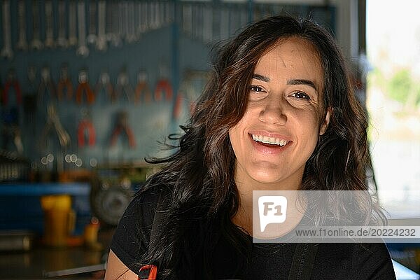 Lächelnde hispanische Mechanikerin in der Werkstatt vor einer Werkbank stehend  eine komplette Werkzeugtafel im Hintergrund mit Bokeh Effekt  traditionelle Männerberufe von Mixed race latino woman