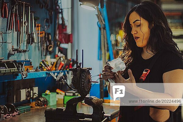 Ein weiblicher Mechaniker in einem Overall in einer Werkstatt reinigt sorgfältig Motorradteile  eine komplette Werkzeugtafel im Hintergrund mit Bokeh Effekt  traditionelle männliche Berufe von Mixed race latino woman