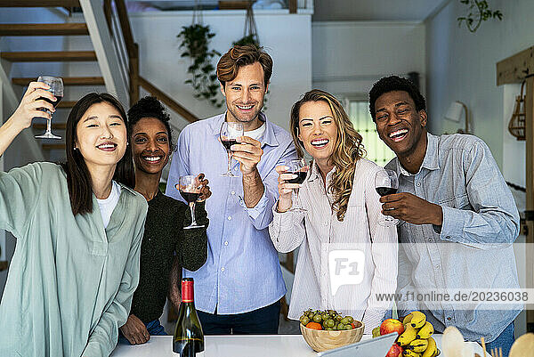 Group of friends looking at the camera toasting with wineglasses standing in kitchen